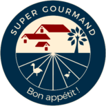 Super Gourmand logo with a circular design. At the top, the text 'RELAIS GOURMET' is arched over a navy blue background. Below, a stylized illustration of a red farmhouse with a white tree stands at the center of a field divided into radial sections, representing a landscape. At the bottom of the circle, it reads 'MAISON GASTRONOMIQUE AU PAYS BASQUE', indicating a gastronomic house in the Basque Country. The logo's background is cream and navy blue, and there are images of two cream-colored ducks at the bottom, one on each side, completing the design.